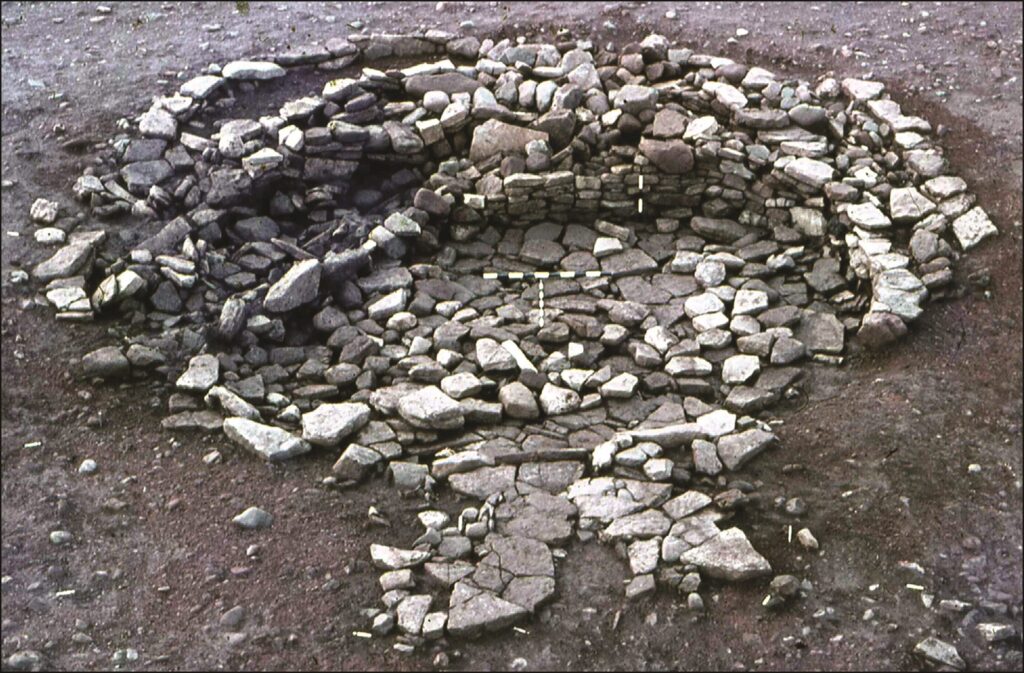 Fotografie eines bronzezeitlichen Rundhauses in Schottland. Das Fundament und die noch erhaltenen Wände bestehen aus großen grau-weißen Steinen. In den Mauern wurden verschiedene Andenken platziert.