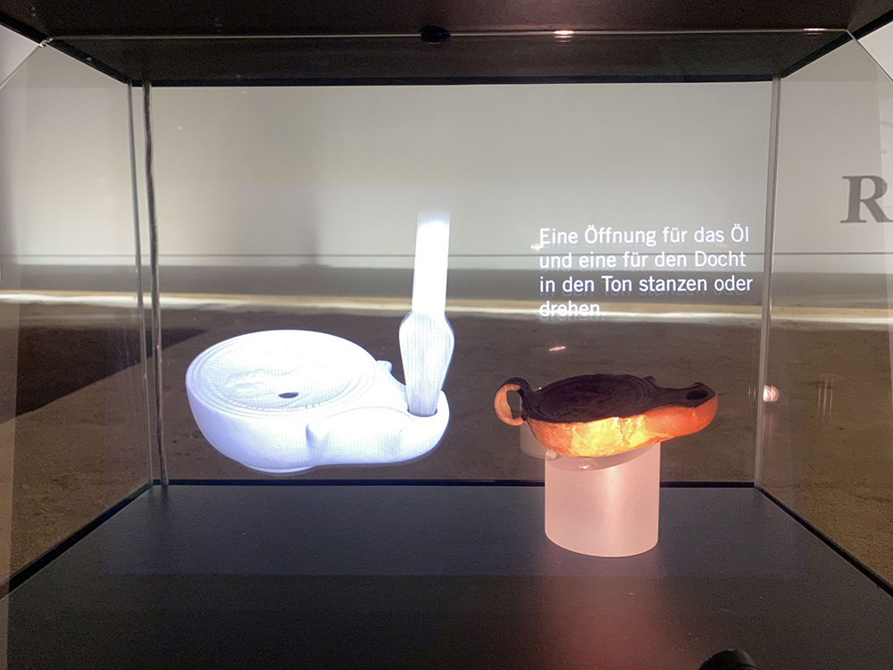 Eine römische Öllampe aus Ton entsteht vor den Augen der Besuchenden, gleich daneben das Original-Exponat im LWL-Archäologie-Museum.